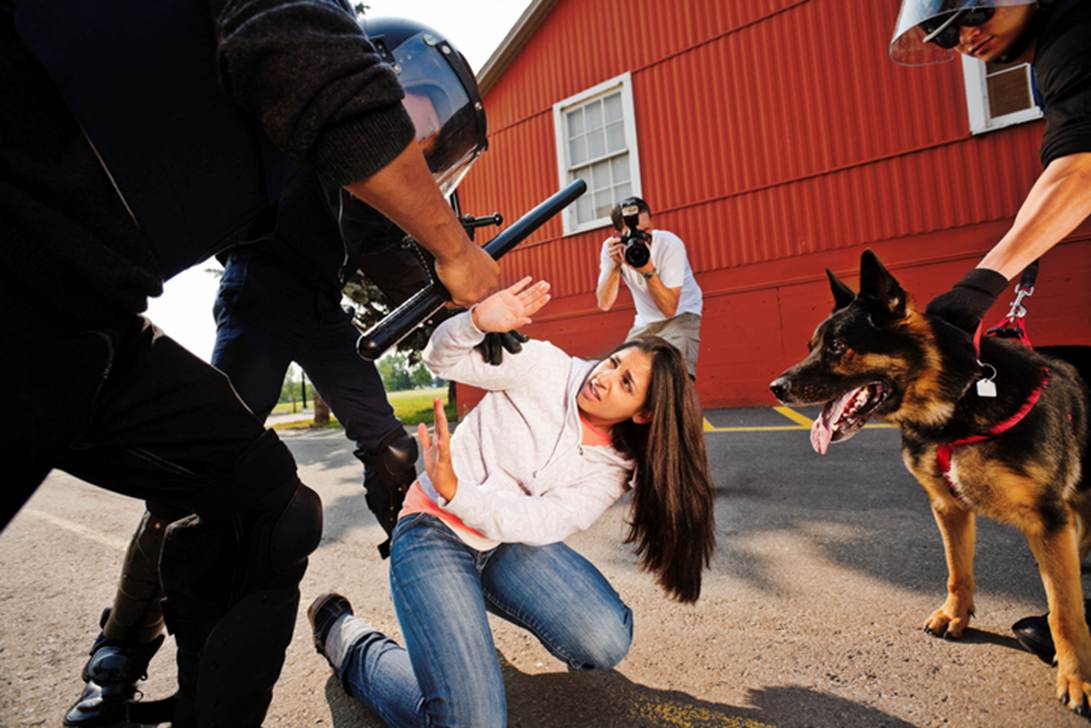 The image shows a woman kneeling with her hands up, surrounded by riot police and a police dog, while a photographer is in the background. It accompanies a discussion on victims becoming unwilling symbols when crushed by publicity.